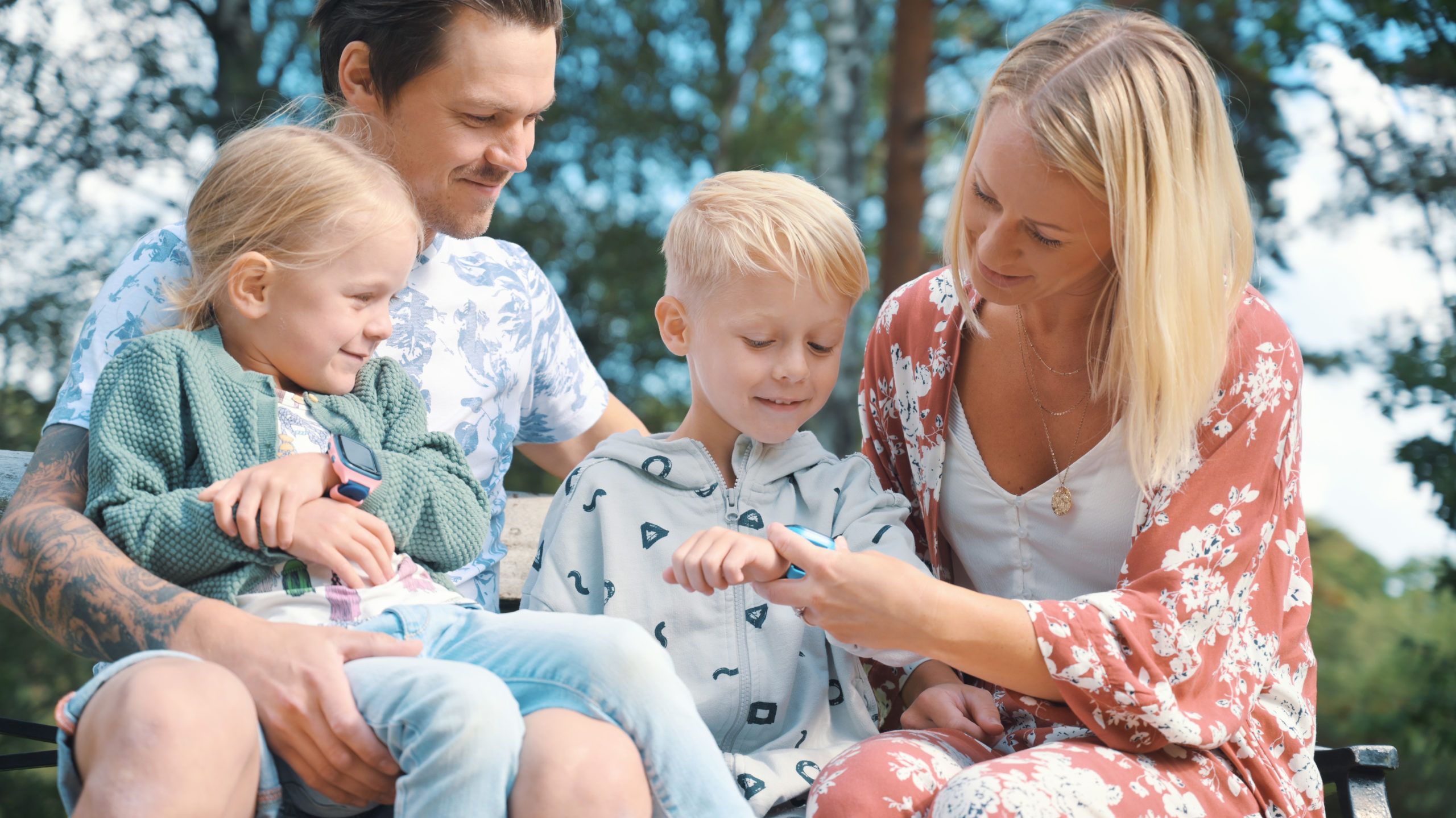 en familie på fire med to barn som har hver som xplora mobilklokke. Fra venstre er det en jente med rosa klokke, en far, en blond gutt med blå klokke og en mor helt til høyre.